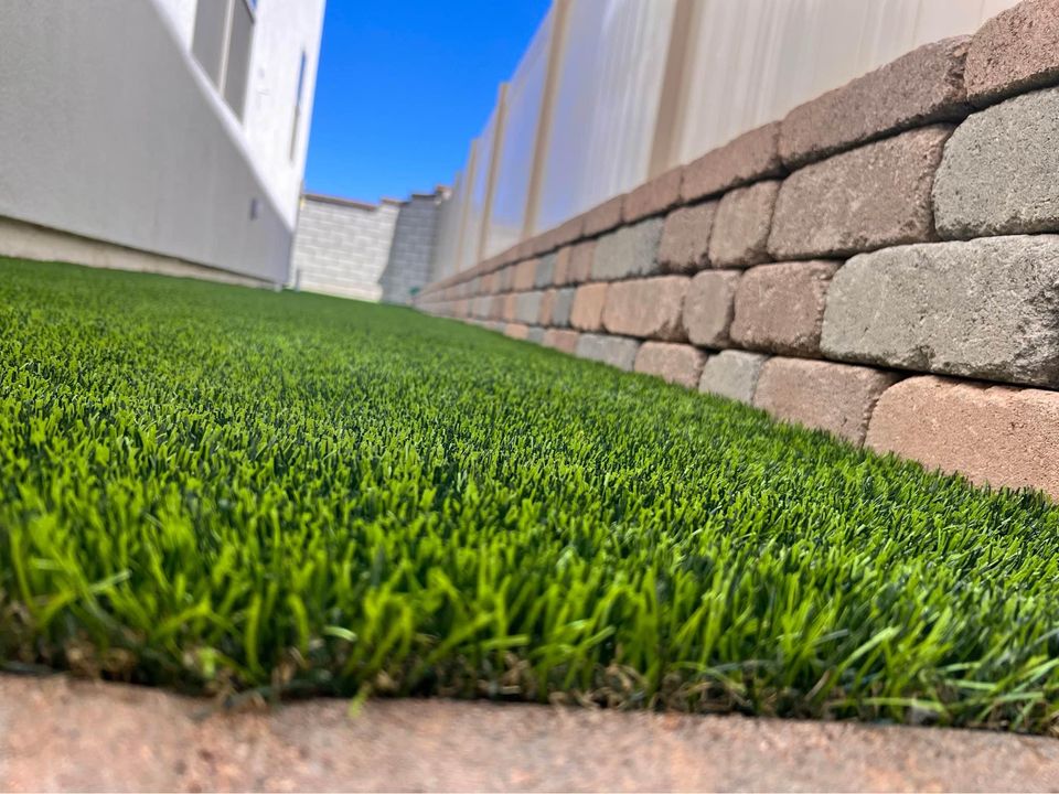Close-up view of a narrow grassy area between a white house and a stone wall. The brick wall has a pattern of alternating beige, gray, and brown stones. The sky is clear and blue, casting bright light on the maintenance-free lawn installed with artificial grass.